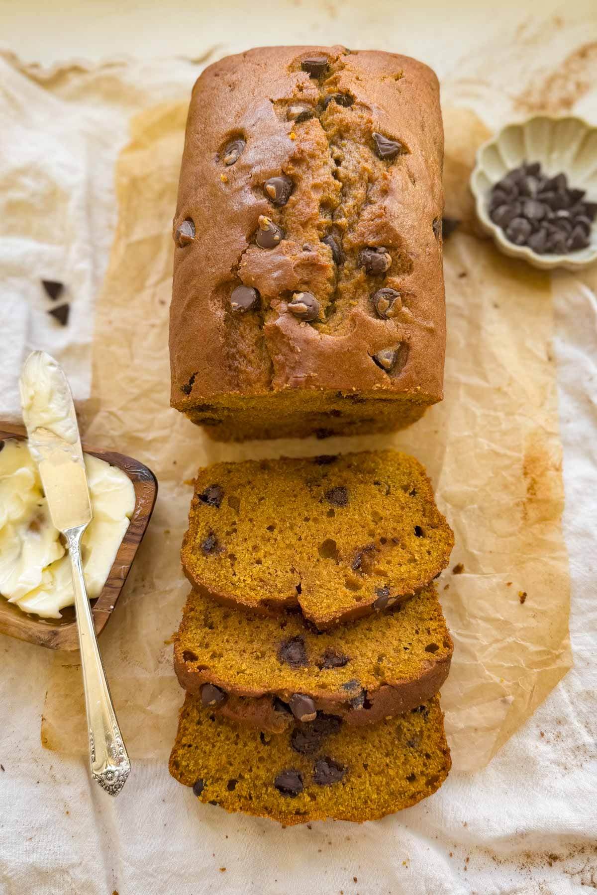 A loaf of chocolate chip pumpkin bread with three slices and a bowl of chocolate chips and butter beside it.