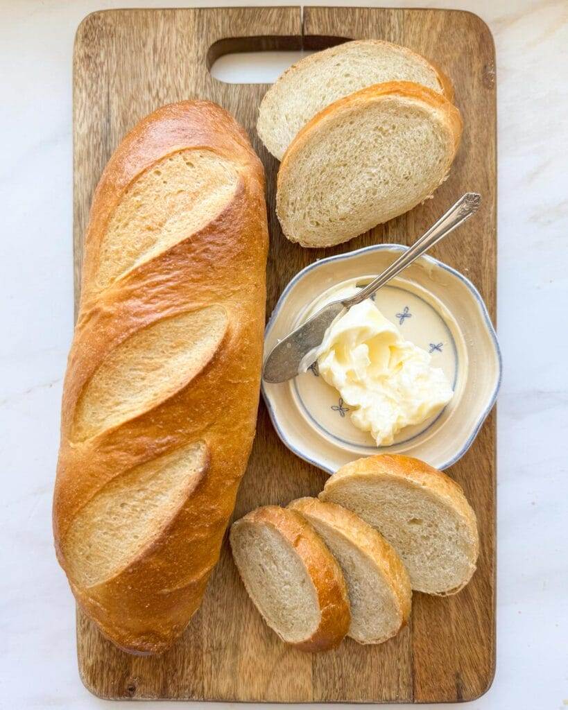 A loaf of french bread on a bread board with sliced french bread and a butter dish.