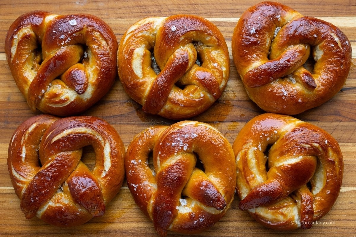 Six baked and salted soft pretzels on a wood cutting board.