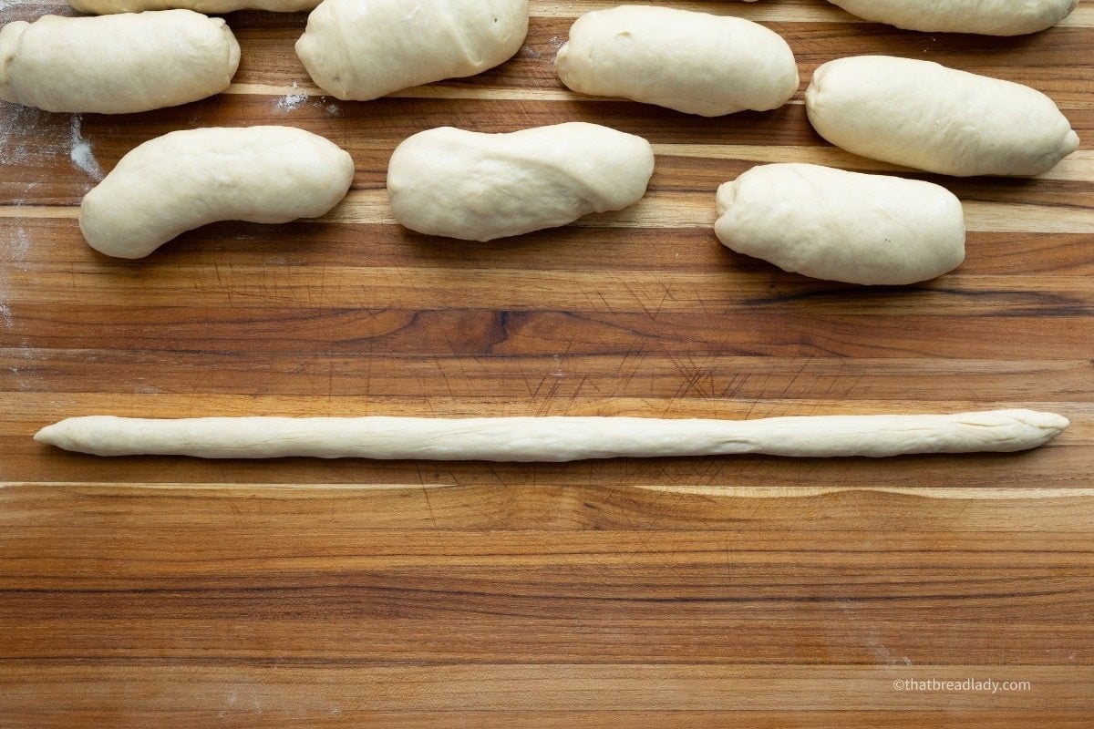 Several rolled up pieces of soft pretzel dough and one long, thin rope of dough, on a wood cutting board.