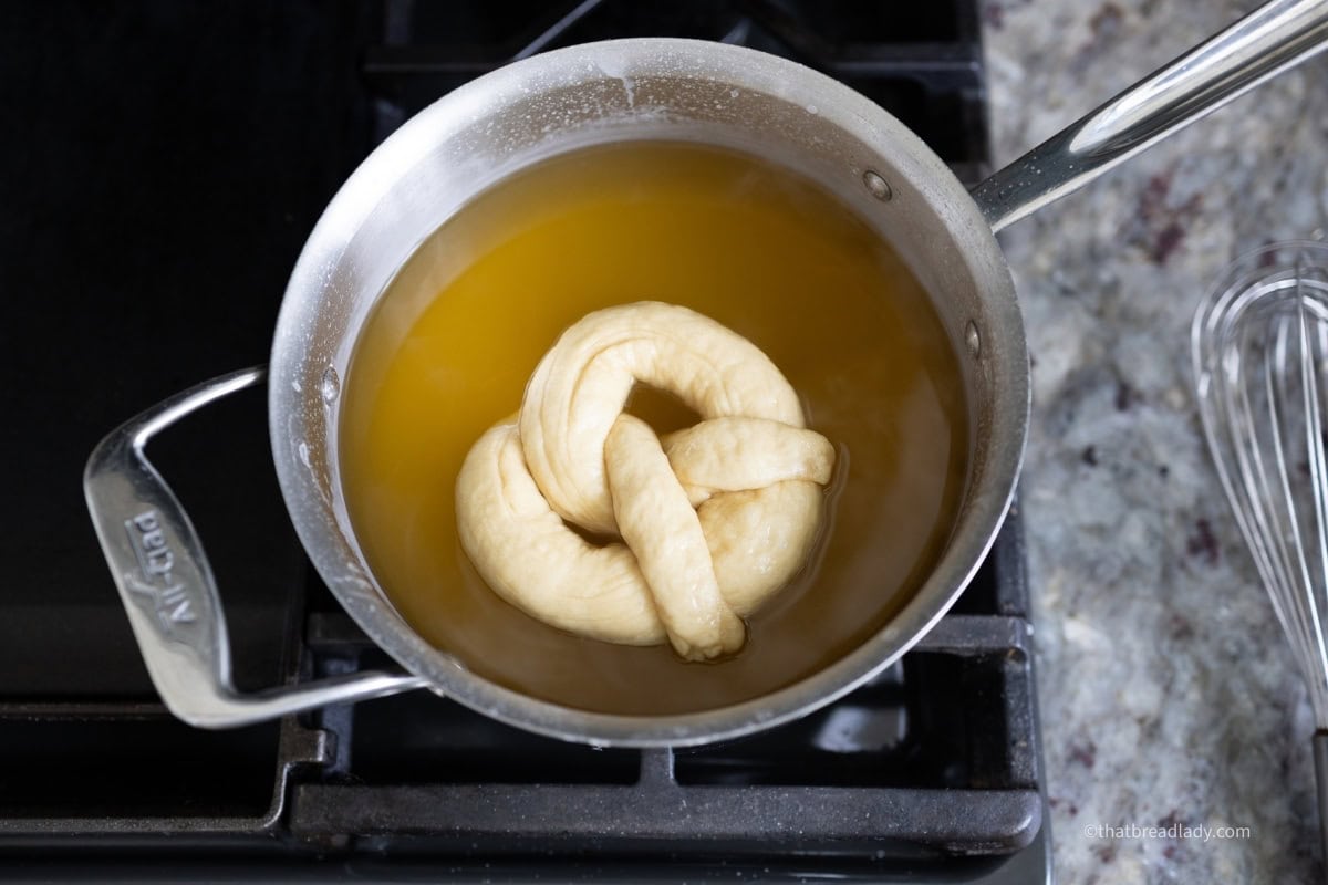 A saucepan filled halfway with water, with a soft pretzel floating in it.