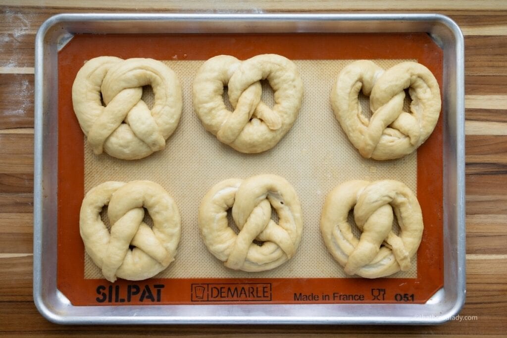 Six soft pretzels resting on a baking sheet, lined with a silicone baking mat.