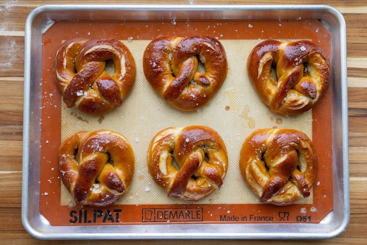 Six baked soft pretzels on a baking sheet lined with a silicone baking mat.