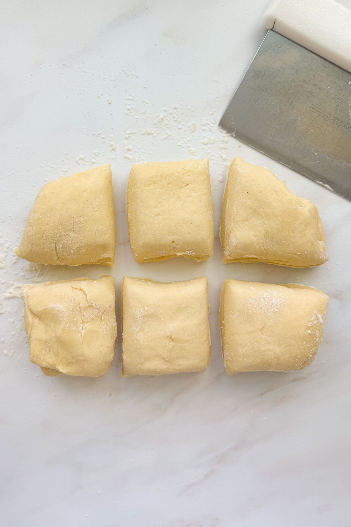 Six pieces of French brioche dough with a bench scraper on a floured marble countertop.