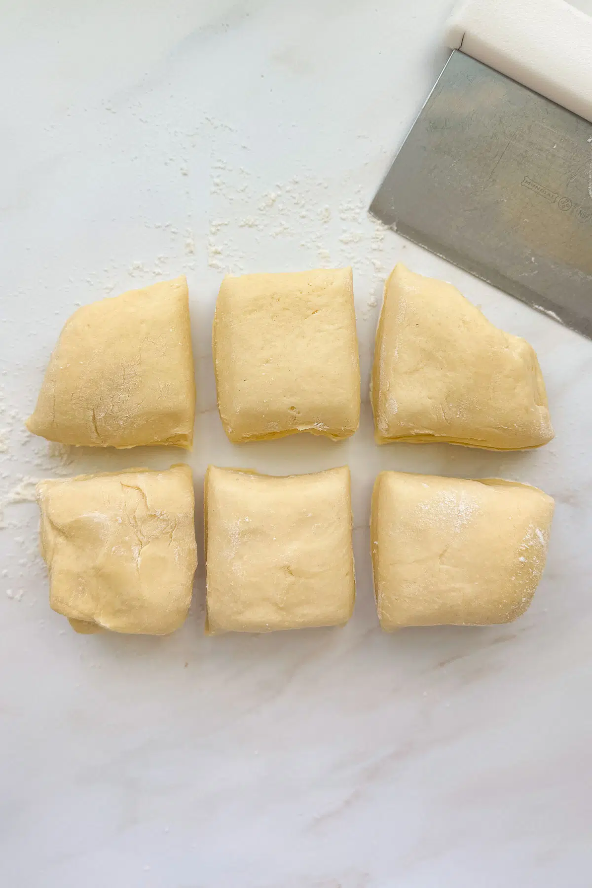 Six pieces of French brioche dough with a bench scraper on a floured marble countertop.
