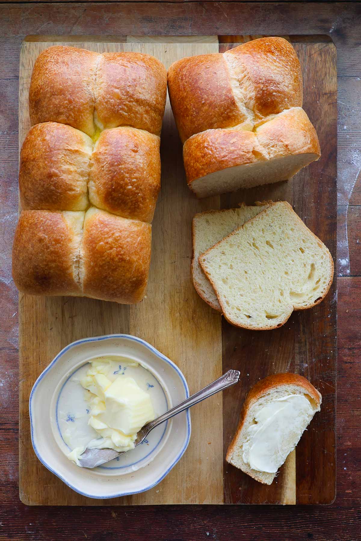 Two loaves of french brioche bread on a cutting board, with one sliced and a butter dish on the board.