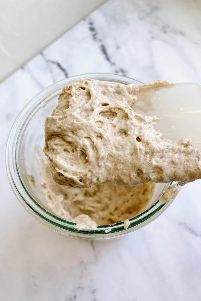 A spatula with sourdough starter, over a glass jar with more sourdough starter.