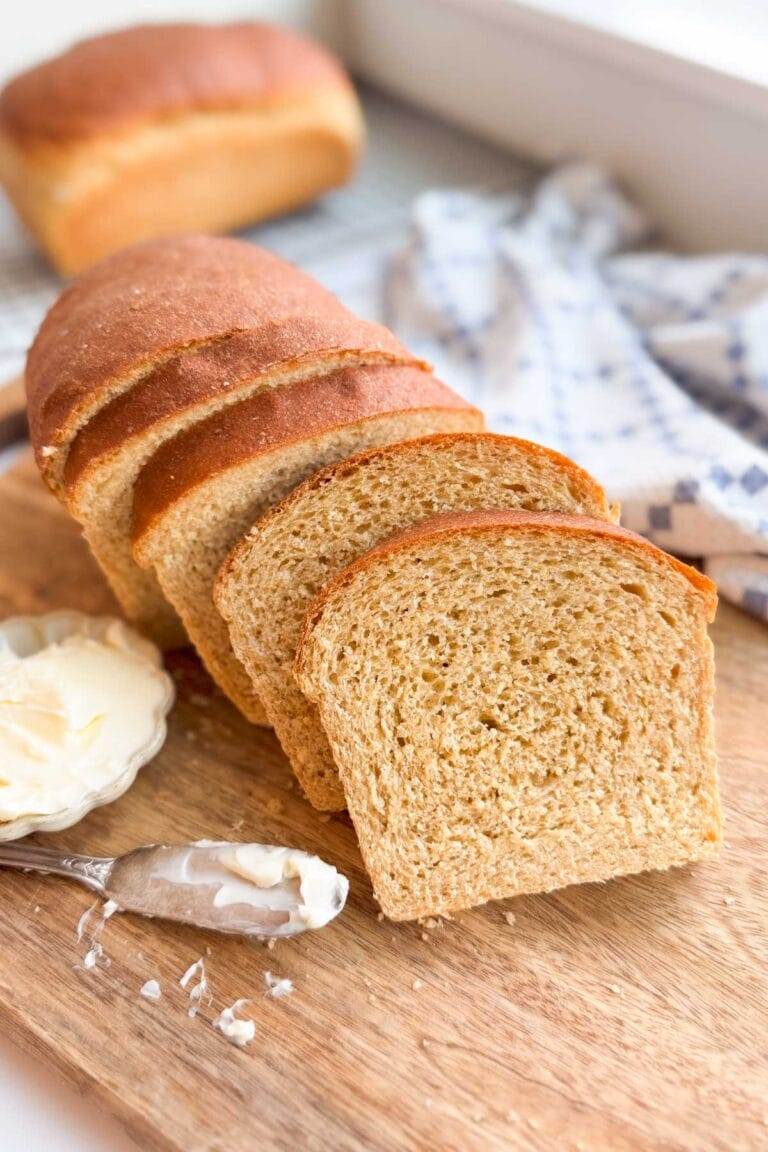 Sliced honey whole wheat sandwich bread on a wood cutting board with a small bowl of butter.