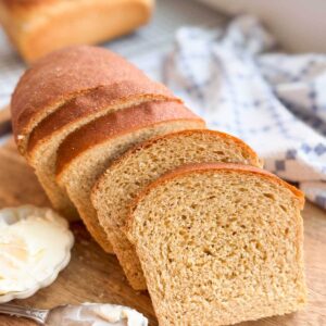 Sliced honey whole wheat sandwich bread on a wood cutting board with a small bowl of butter.