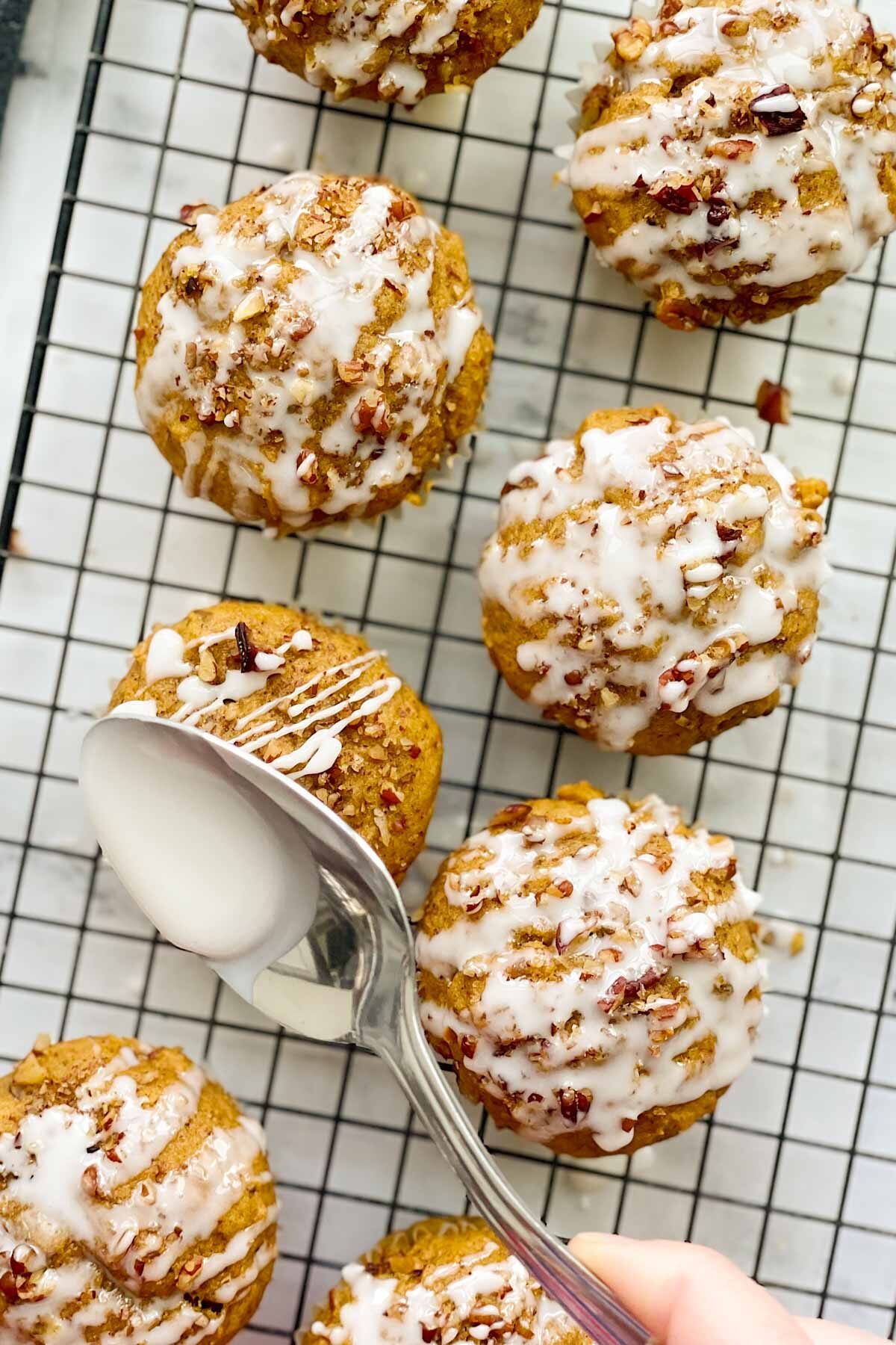 Several pumpkin sourdough muffins on a cooling rack, with icing on top and a hand holding a spoon with icing, adding more to the top of one muffin.