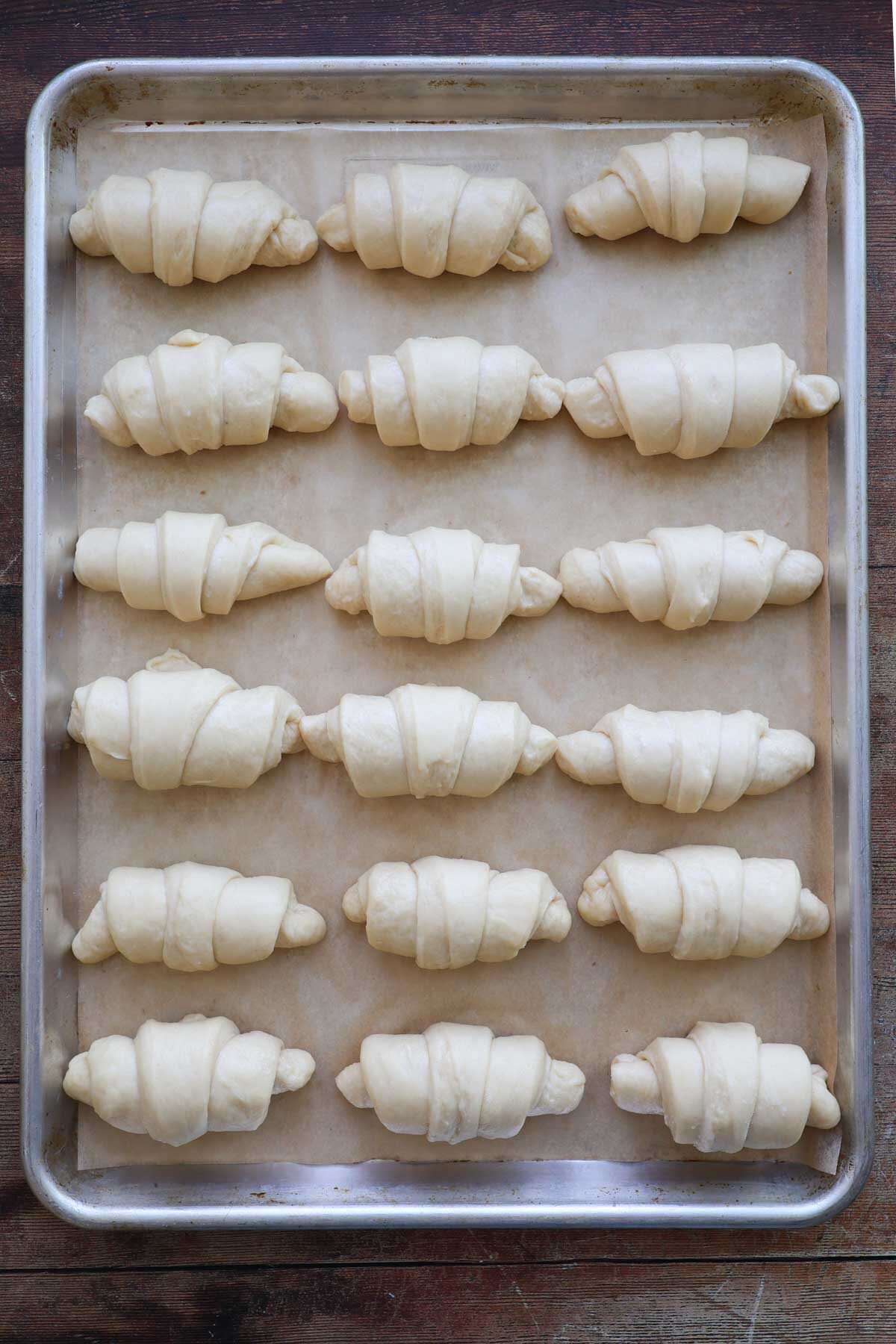 A sheet pan lined with parchment paper, and 18 unrisen, unbaked, crescent rolls on it with 6 rows of rolls and three rolls across.