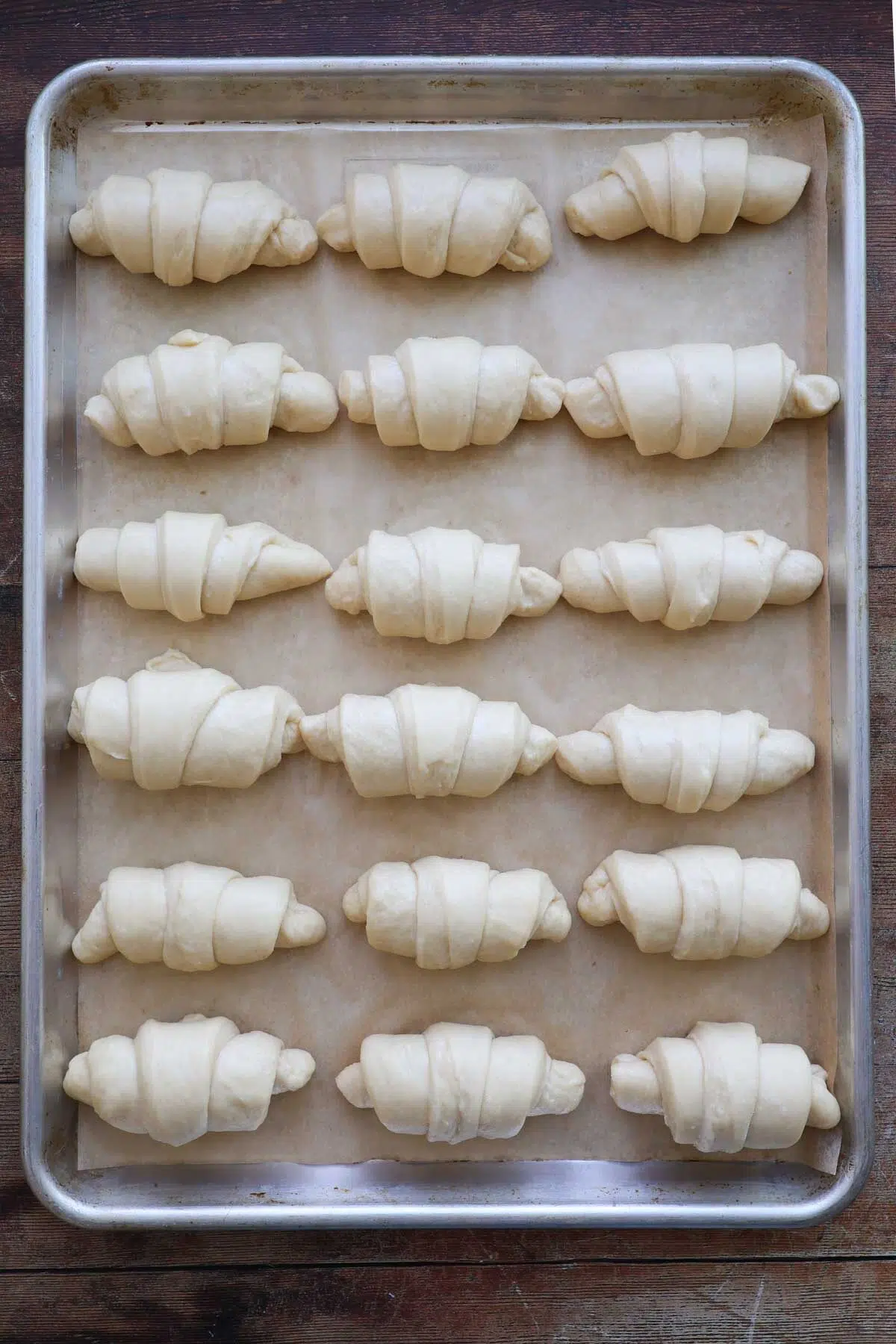 A sheet pan lined with parchment paper, and 18 unrisen, unbaked, crescent rolls on it with 6 rows of rolls and three rolls across.