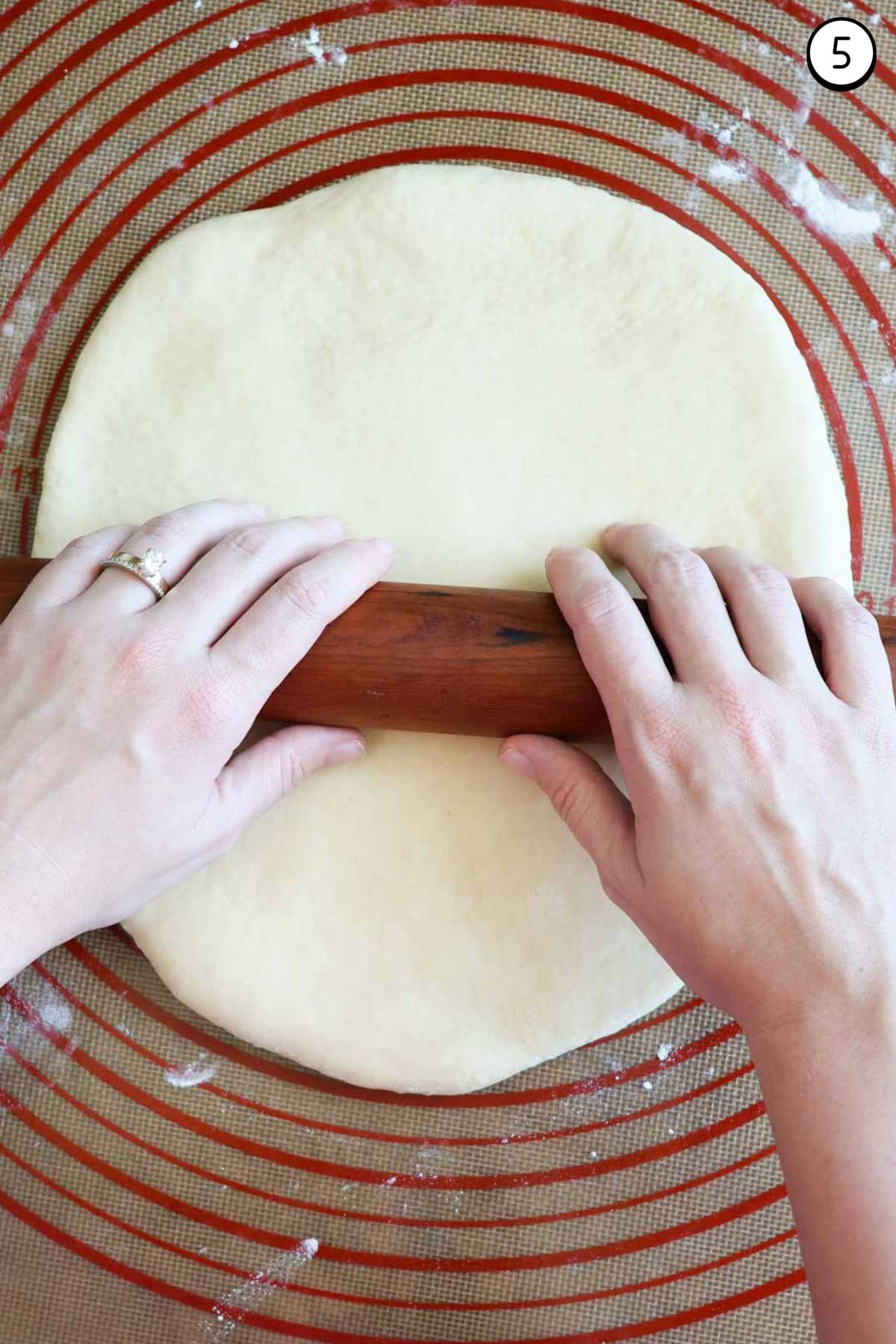 Two hands on a wood rolling pin, rolling out crescent roll dough on top of a silicone rolling mat.