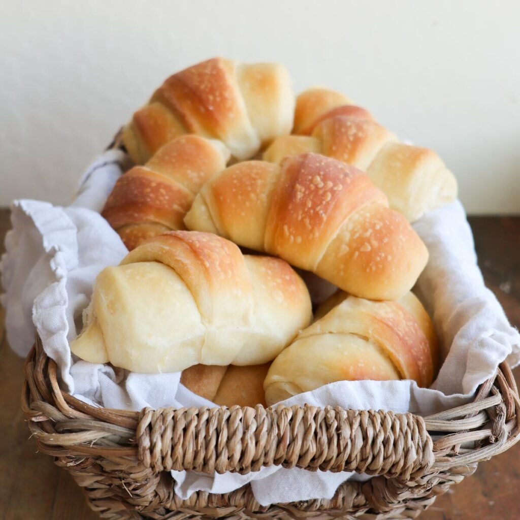 A basket lined with a linen napkin and full of fluffy homemade crescent rolls.