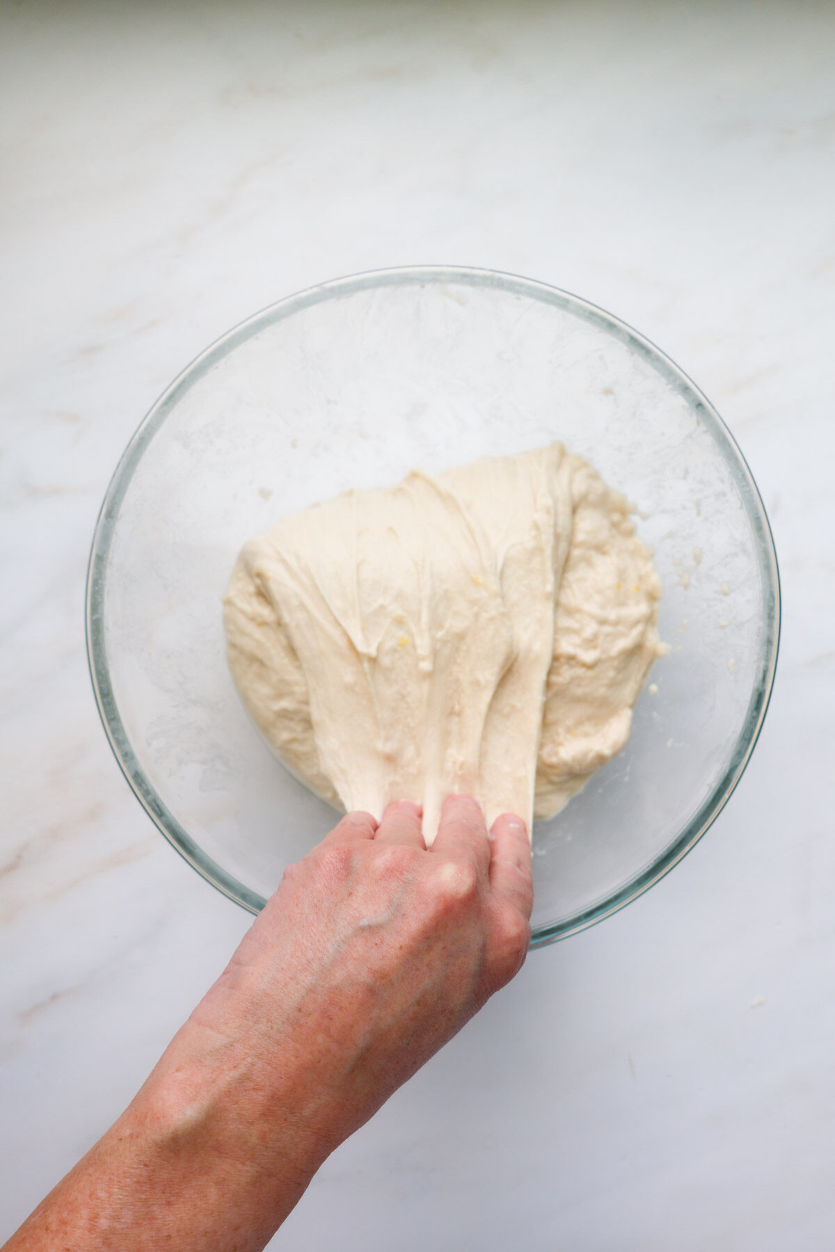 A glass bowl with dough in it, with a hand folding the top portion of the dough over.