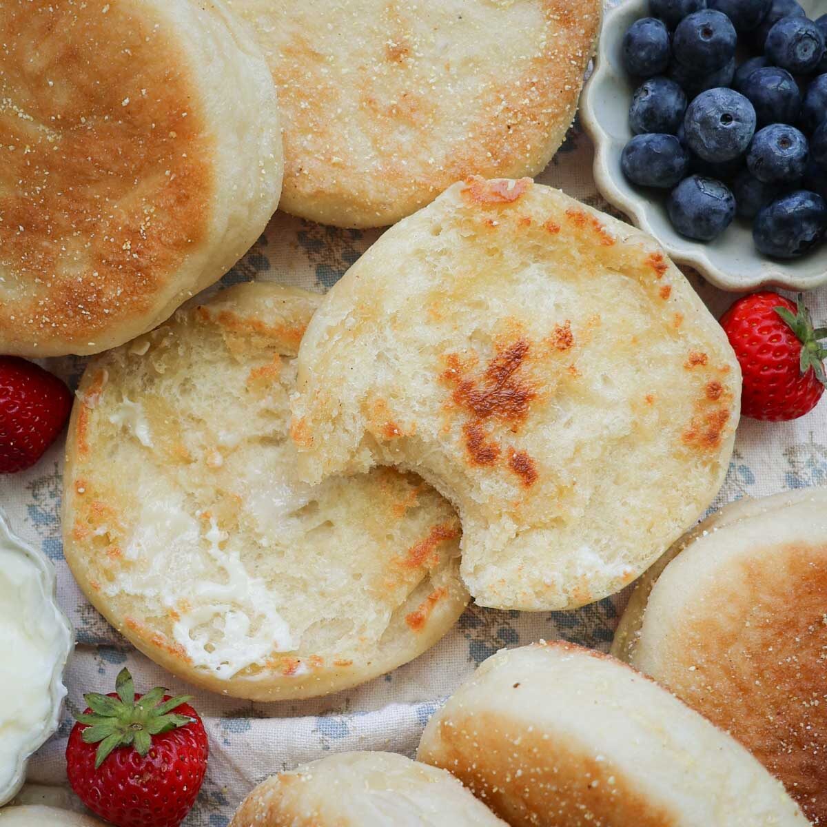 A sourdough English muffin split in half, toasted, buttered, with one bite taken, surrounded by more sourdough English muffins, and blueberries and strawberries.