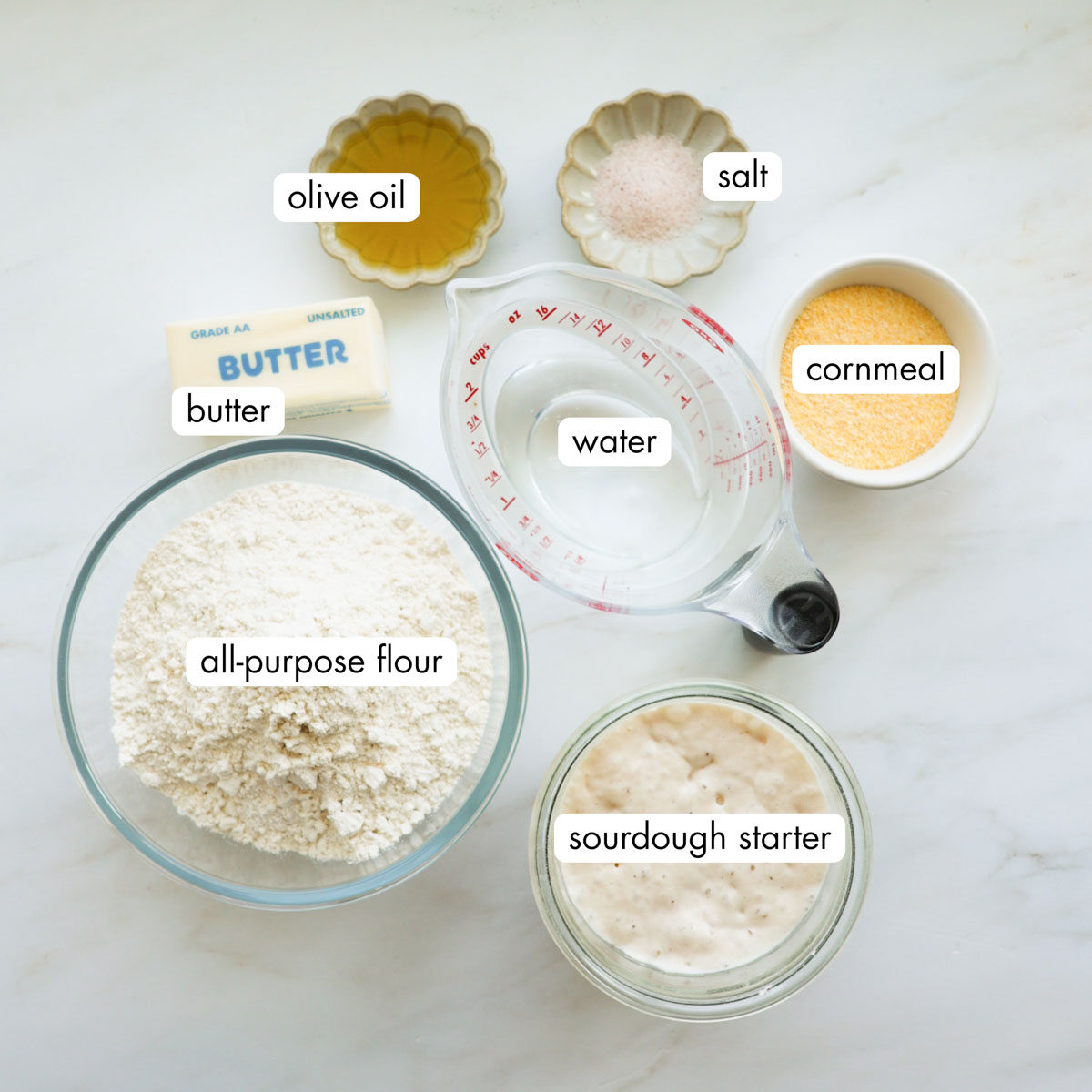 All the ingredients for sourdoughough English muffins measured out, including all-purpose flour, butter, water, olive oil, salt, cornmeal, and sourdough starter.
