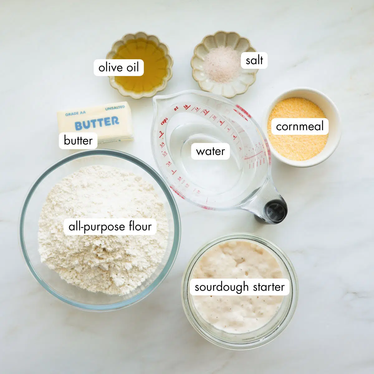 All the ingredients for sourdoughough English muffins measured out, including all-purpose flour, butter, water, olive oil, salt, cornmeal, and sourdough starter.