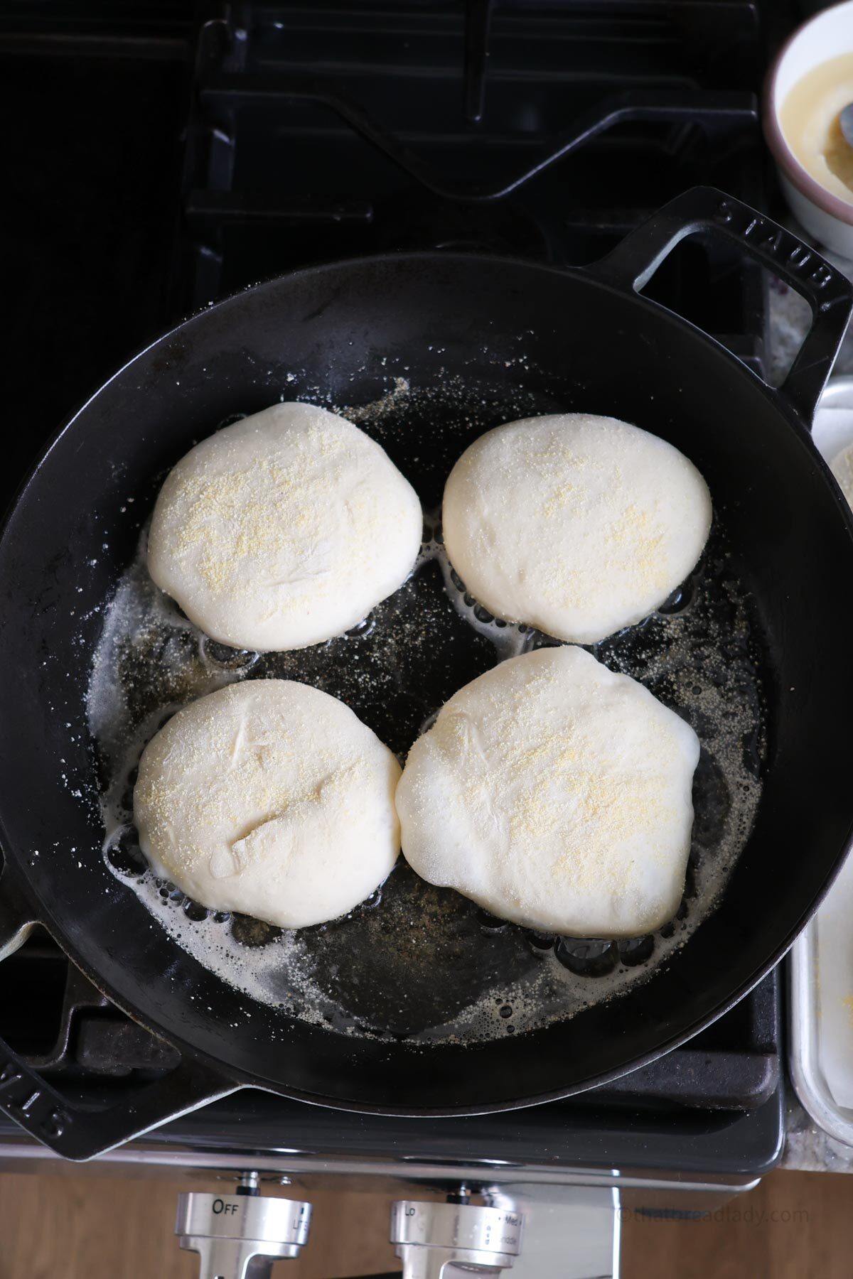 Four english muffins cooking in butter in a cast iron skillet on the stove top.
