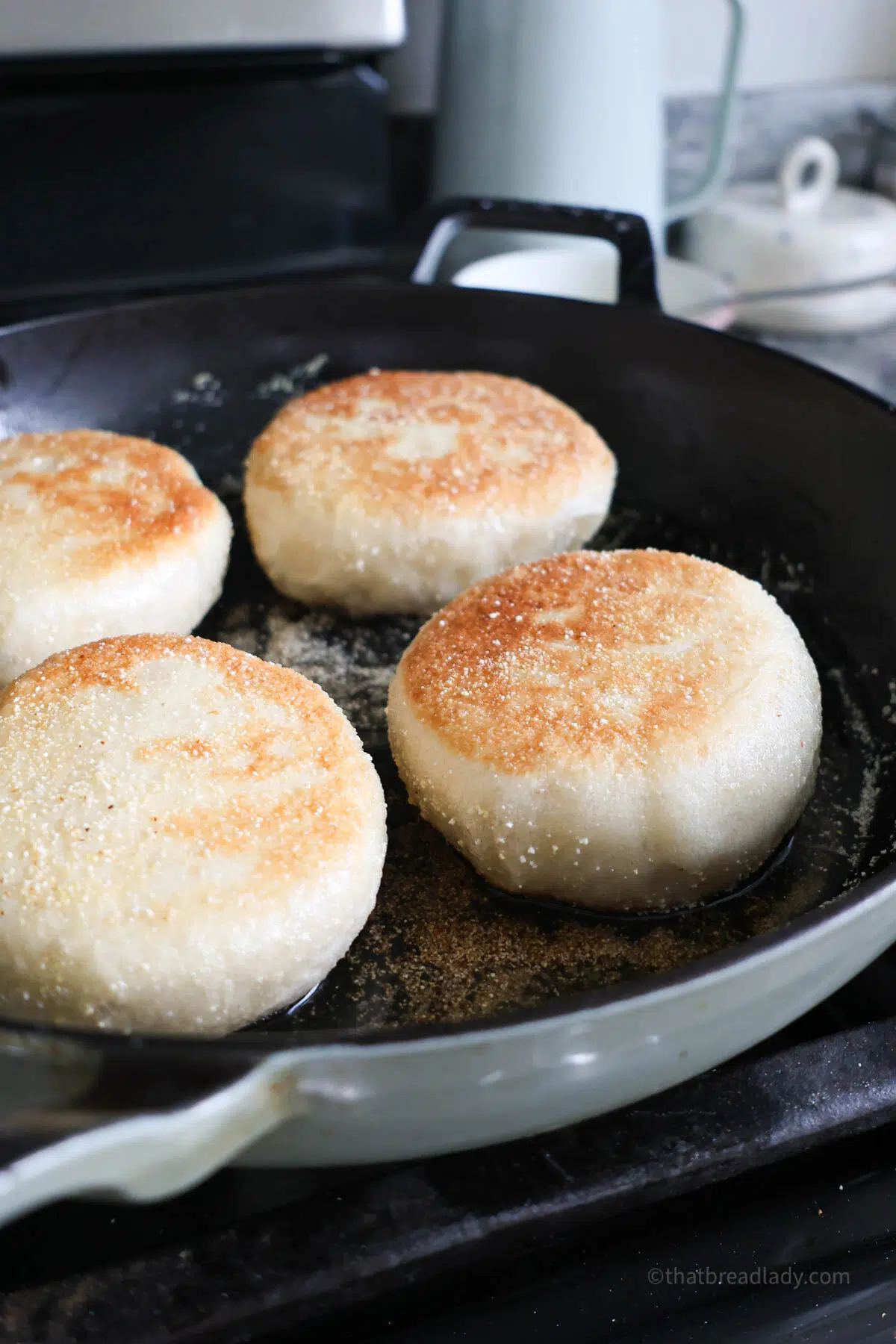 Four English muffins cooking in a cast iron skillet with toasted tops.
