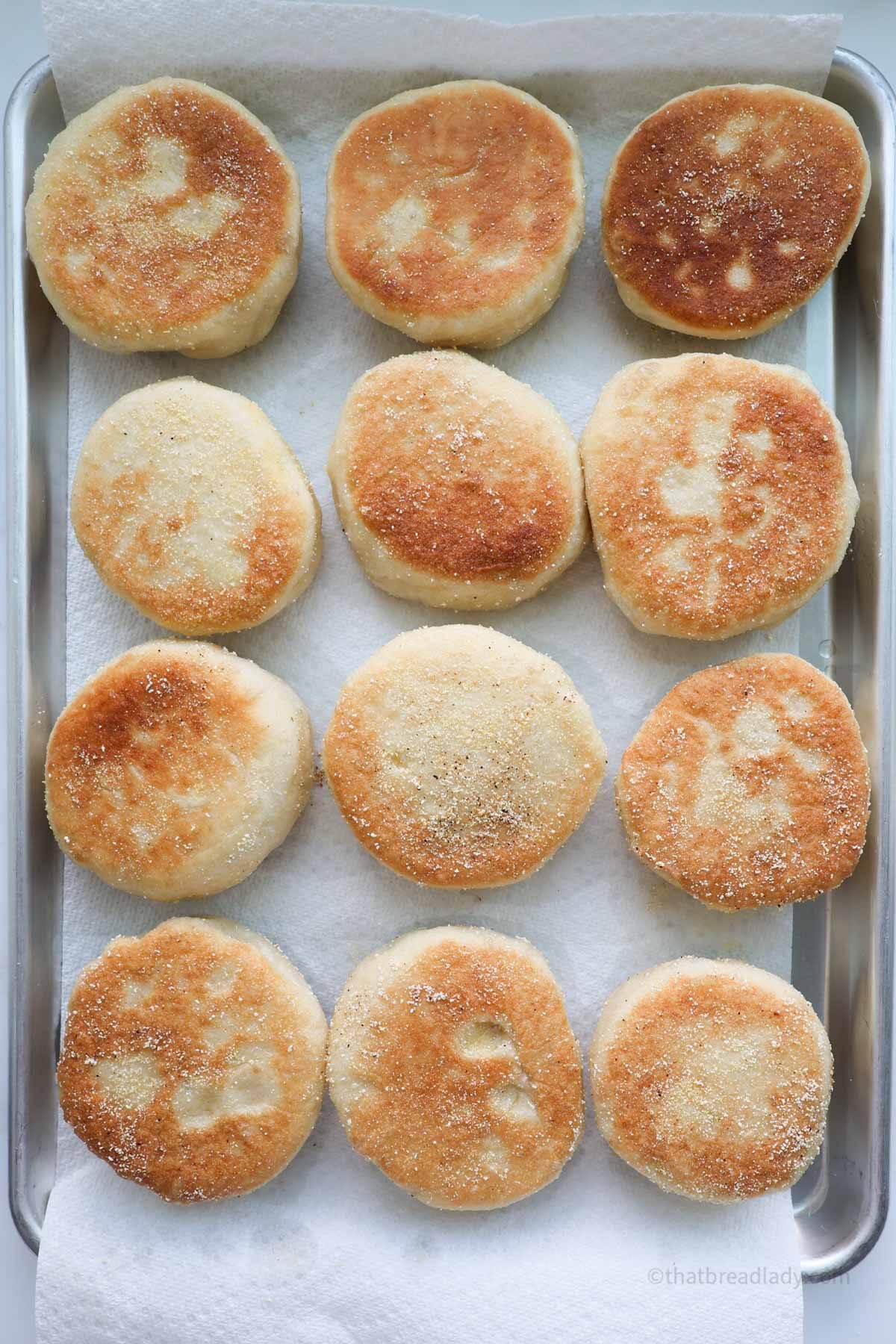 A baking sheet lined with paper towels and 12 cooked English muffins on top.