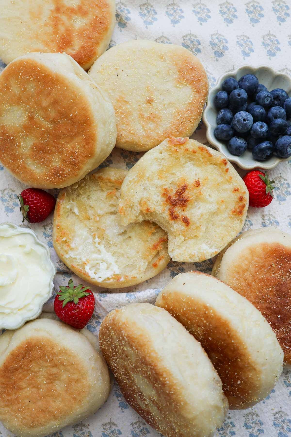 A sourdough English muffin that has been split apart, toasted, and buttered, with one bit taken out of one of them. They are surrounded by many more english muffins plus blueberries, strawberries, and a small bowl of butter.