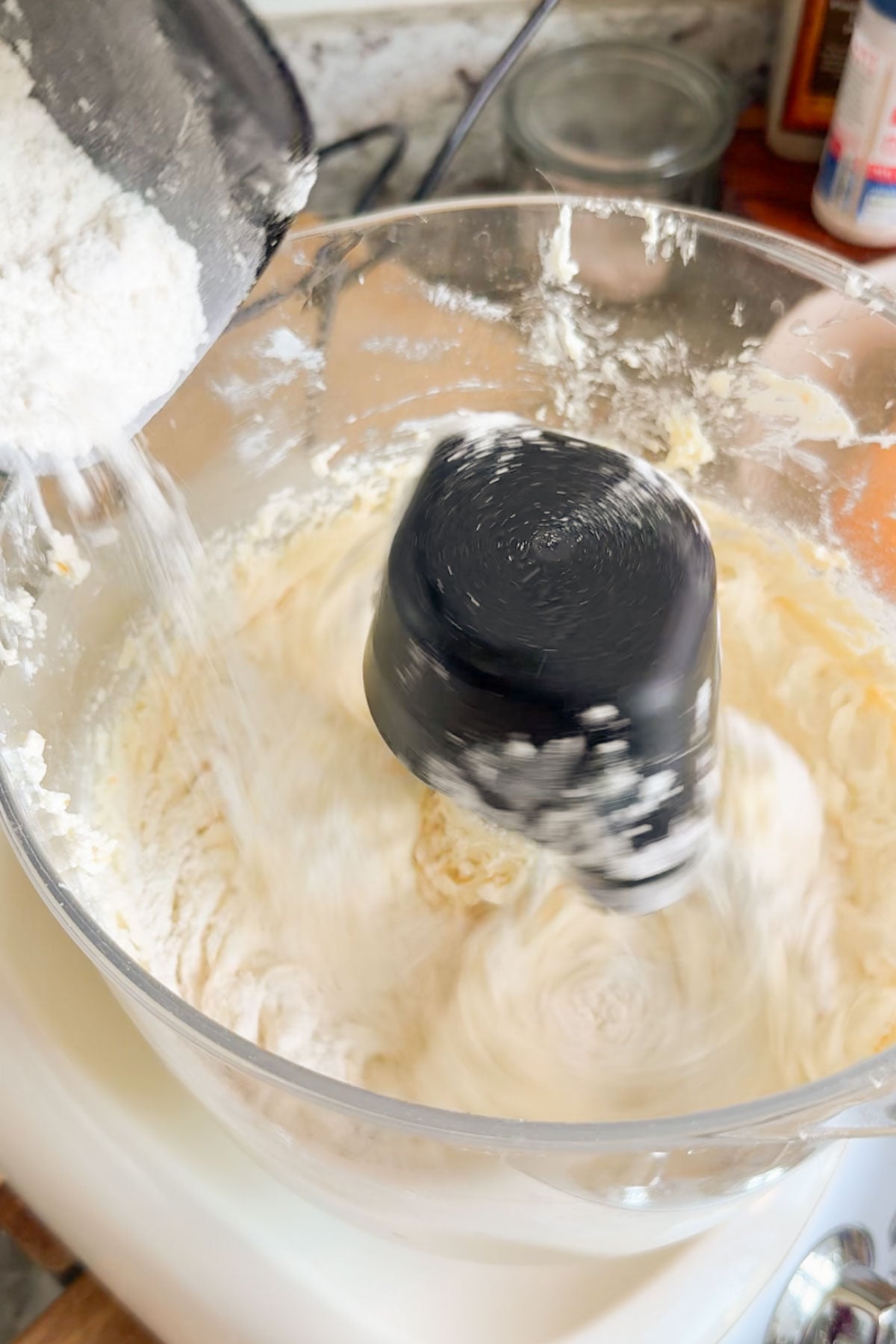 A stand mixer bowl with the mixture of orange pound cake being mixed with a wire whisk and flour being poured into the bowl.