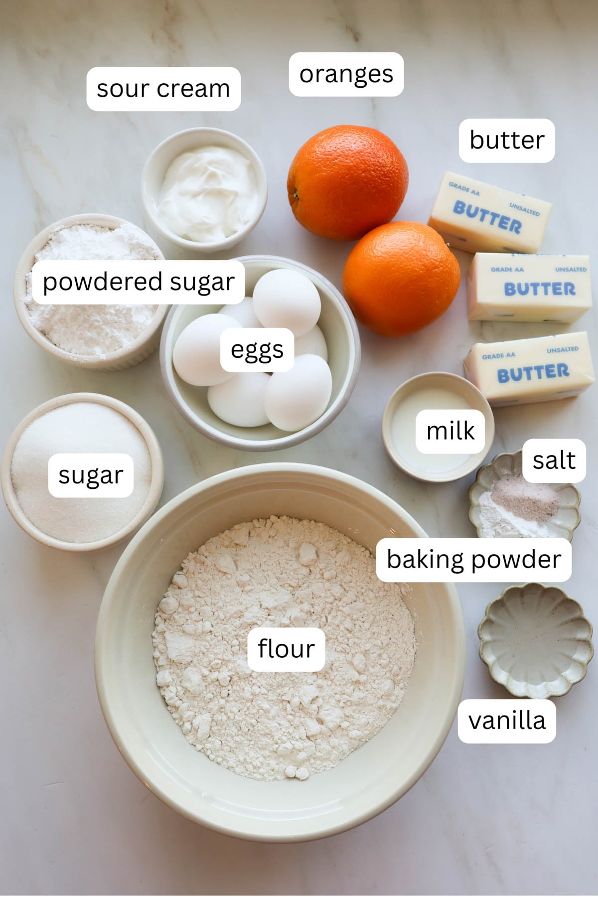 All the ingredients for orange pound cake including butter, sugar, oranges, eggs, vanilla, salt, baking powder, sour cream, flour, powdered sugar and milk.