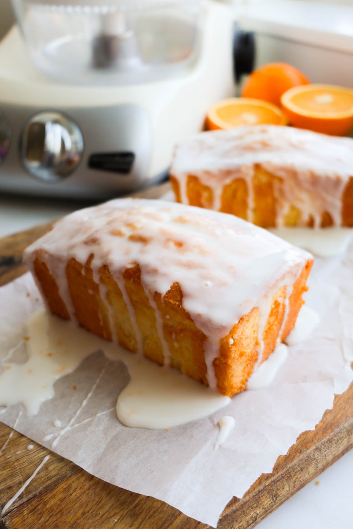 A loaf of orange pound cake, iced with orange zest glaze, resting on a piece of parchment on top of a wood cutting board.