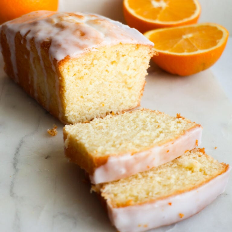 A sliced loaf of orange pound cake, with sliced oranges behind it.