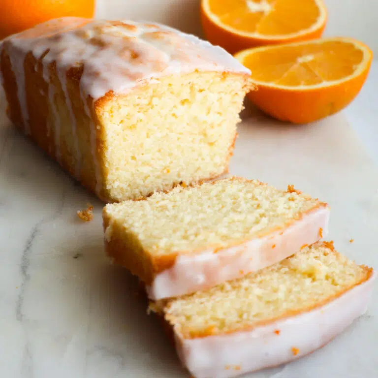 A sliced loaf of orange pound cake, with sliced oranges behind it.