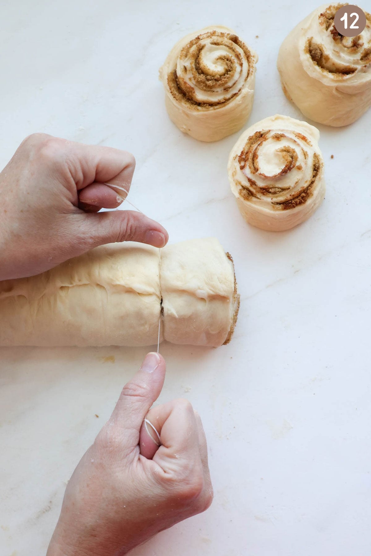 Two hands holding a piece of floss, cutting a cinnamon roll out of a long rolled up piece of dough, with three already cut cinnamon rolls off to the side.