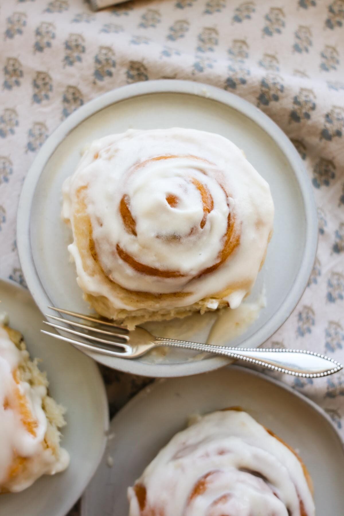 A frosted cinnamon roll on a plate with a fork and more plated cinnamon rolls next to it.