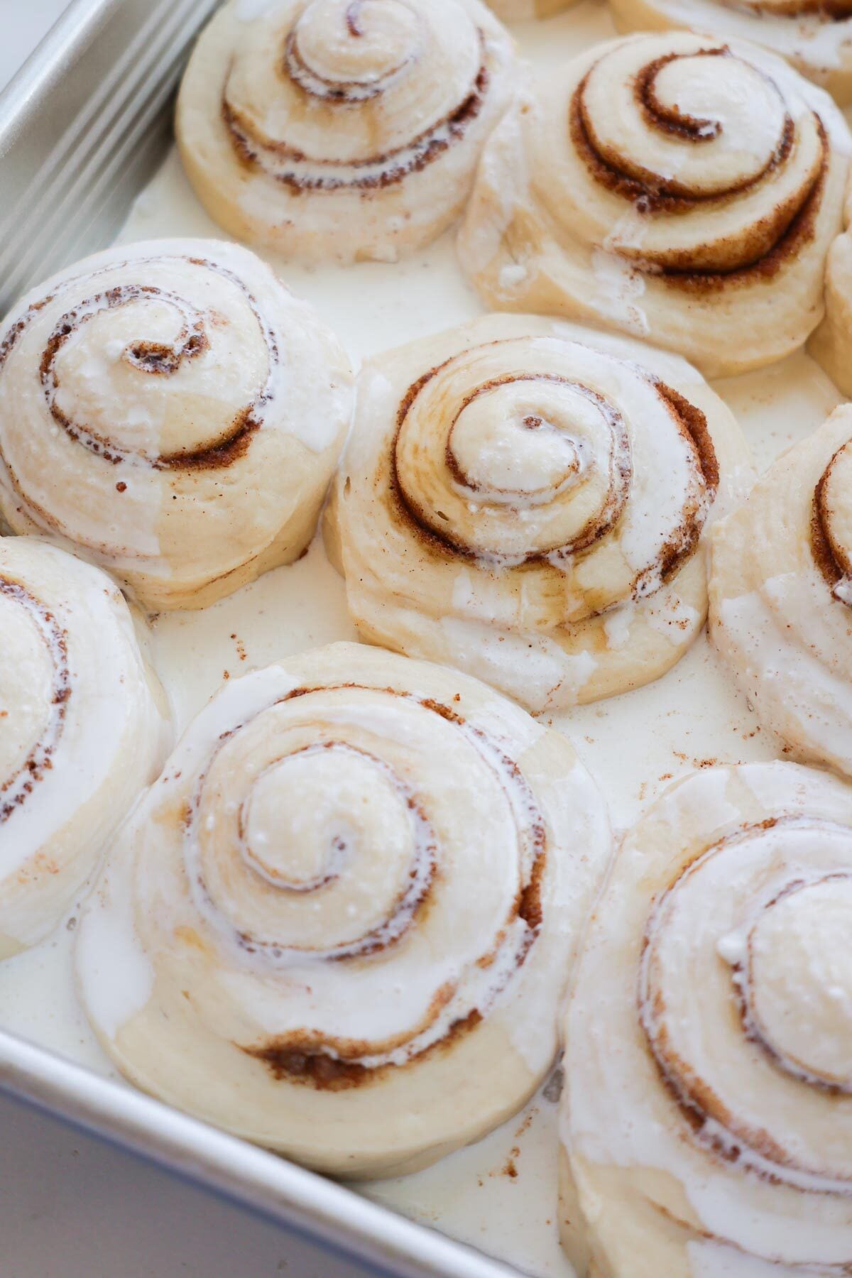 A pan of unbaked cinnamon rolls with heavy cream in the bottom of the pan and some that has been poured over them.