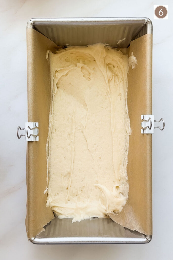 A bread pan lined with parchment paper held in place with two white clips and bread batter spread in the bottom of the pan.