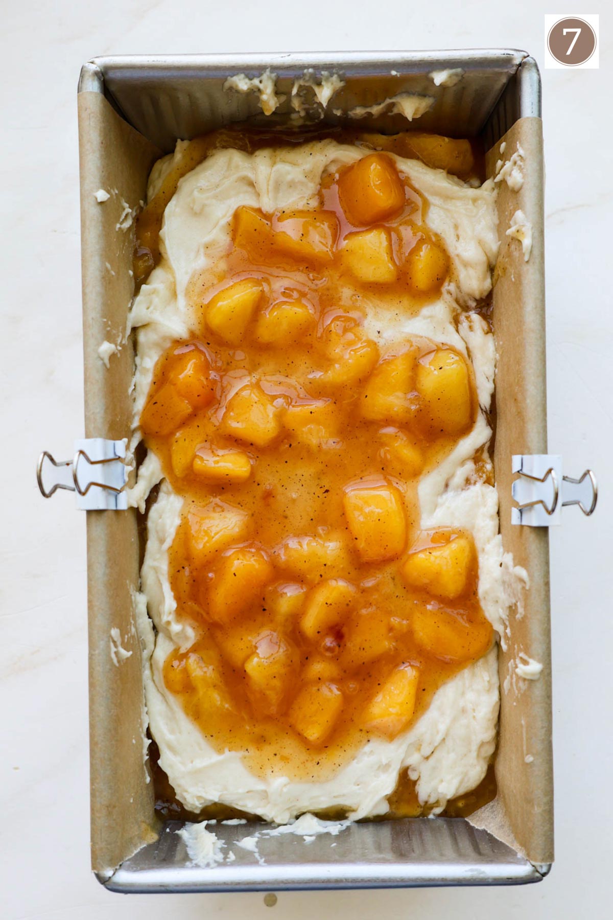 A bread pan lined with parchment paper held in place with two white clips and filled with bread batter and peaches on top.