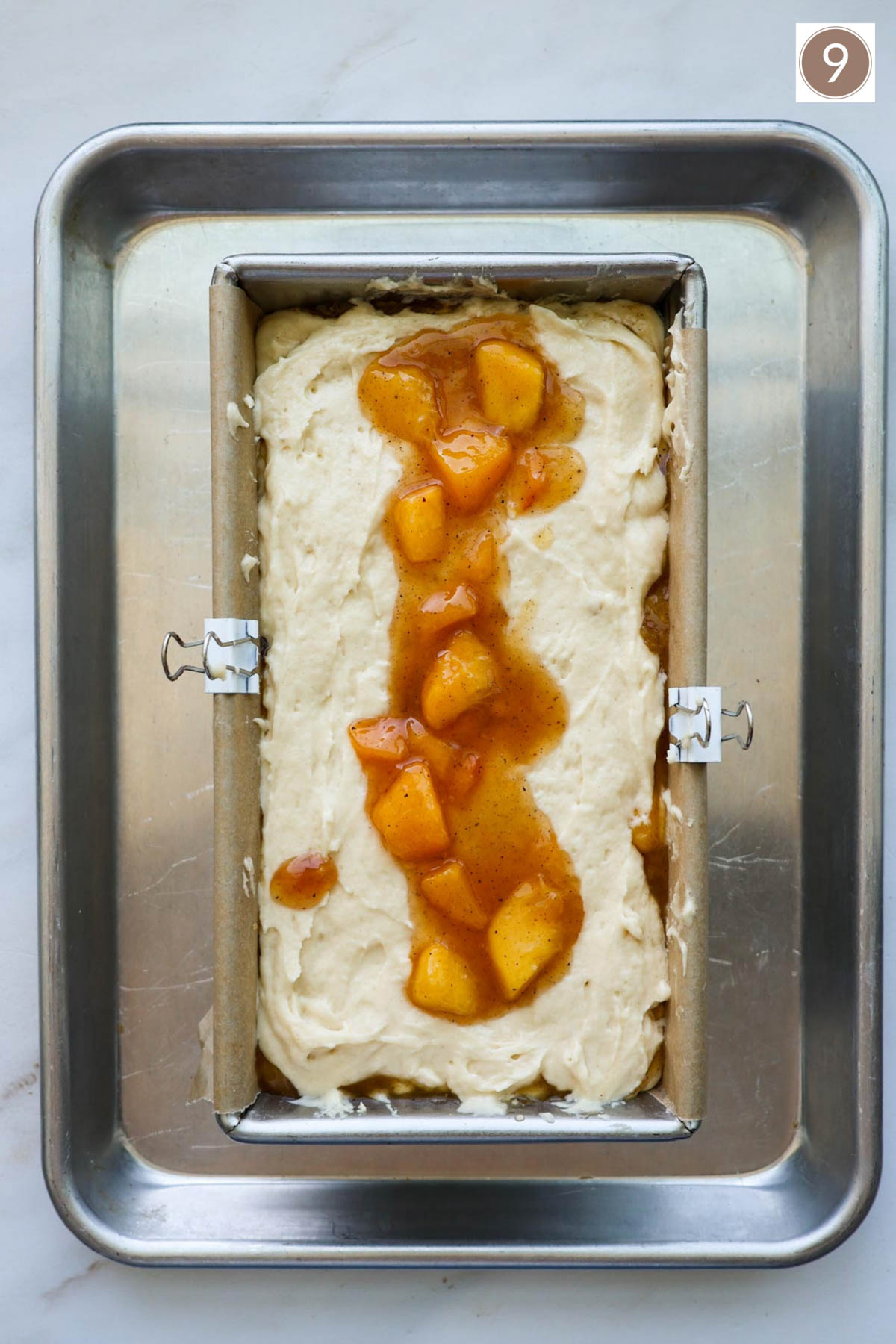A bread pan lined with parchment paper held in place with two white clips and filled with bread batter and peaches on top and resting on top of a baking sheet.