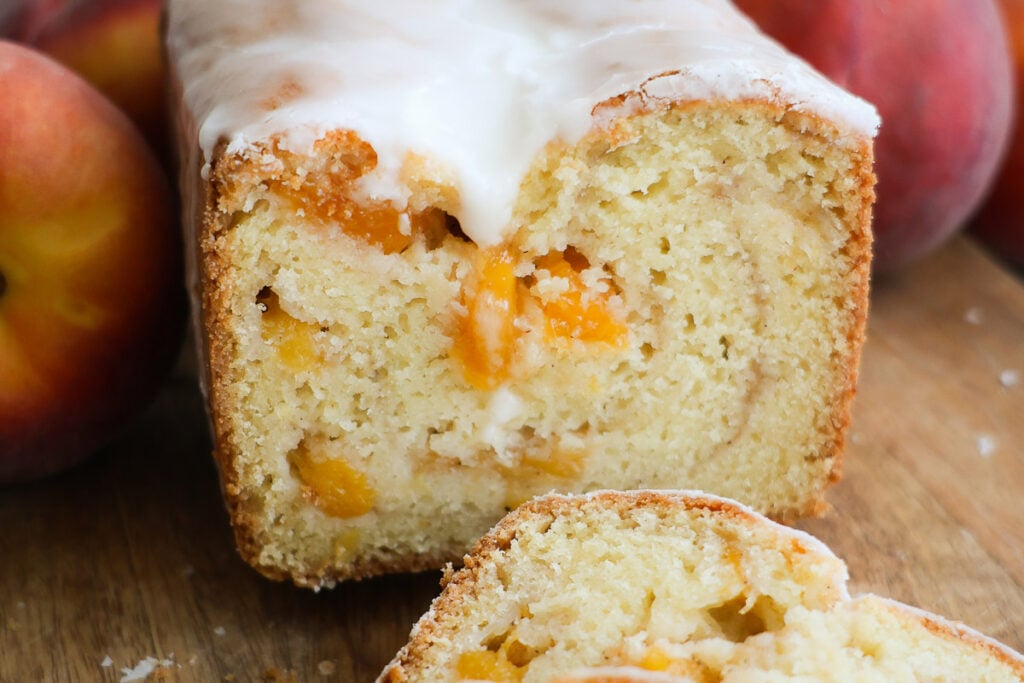 A cross-section of a loaf of peach bread and whole peaches on either side of it.