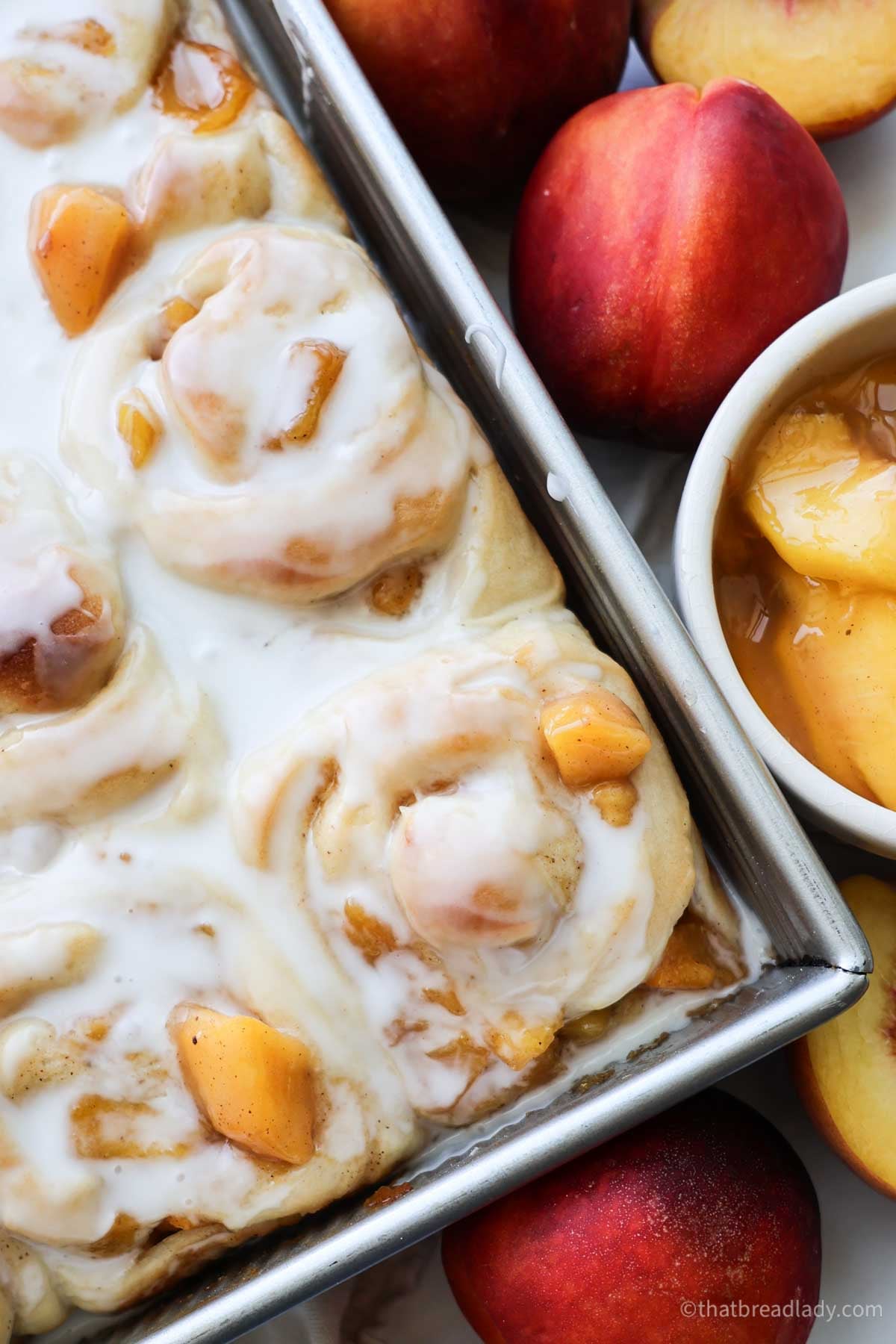 Frosted peach cinnamon rolls, in a metal pan, with peaches around the pan.