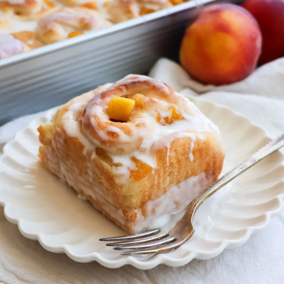A peach cinnamon roll on a white plate with a peach in the background.