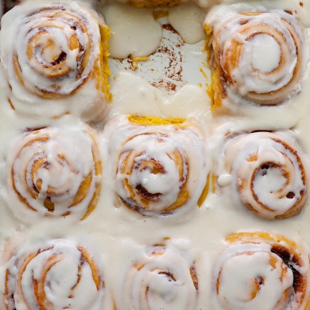 Pumpkin cinnamon rolls, frosted, in a pan, with one missing.