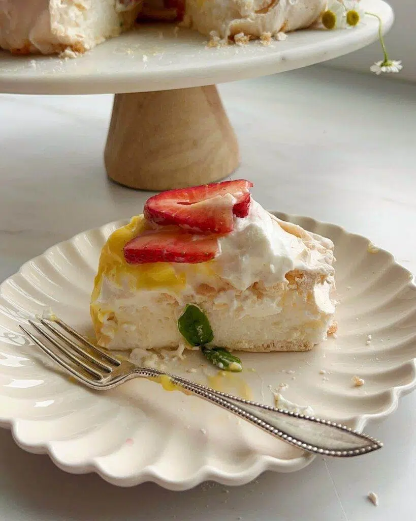 A slice of pavlova, on a dessert plate with a fork, and strawberries and whipped cream on top of the pavlova slice.