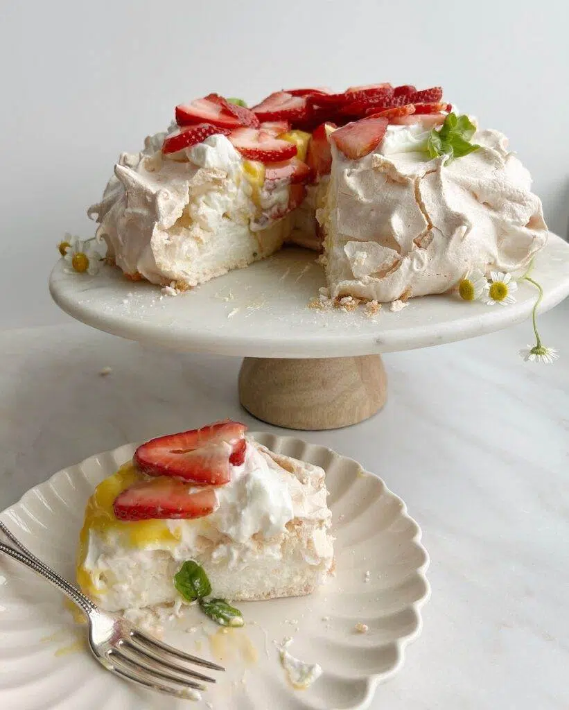 A baked pavlova on a cake stand, garnished with whipped cream, sliced strawberries, a sprig of basil and wild flowers, with one slice on a dessert plate on the counter.