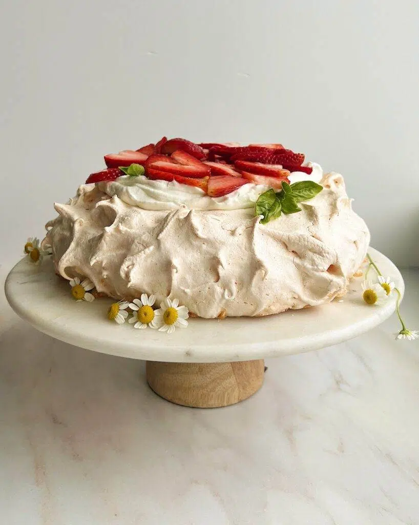 A baked pavlova on a cake stand, garnished with whipped cream, sliced strawberries, sprigs of basil, and wild flowers.