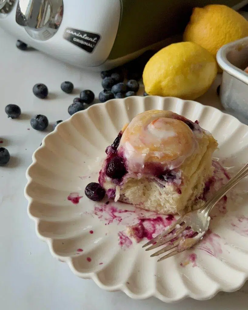 A lemon blueberry sweet rolls on a dessert plate, with a fork, and lemons and blueberries on the counter.