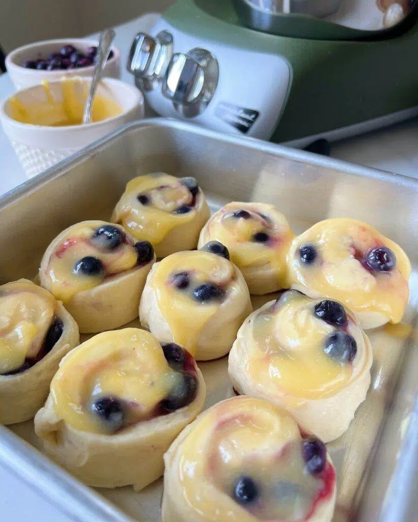 A metal baking pan with 9 lemon blueberry sweet rolls in it, with lemon curd and blueberries on top of the rolls, a mixer, and bowls of lemon curd and blueberries.
