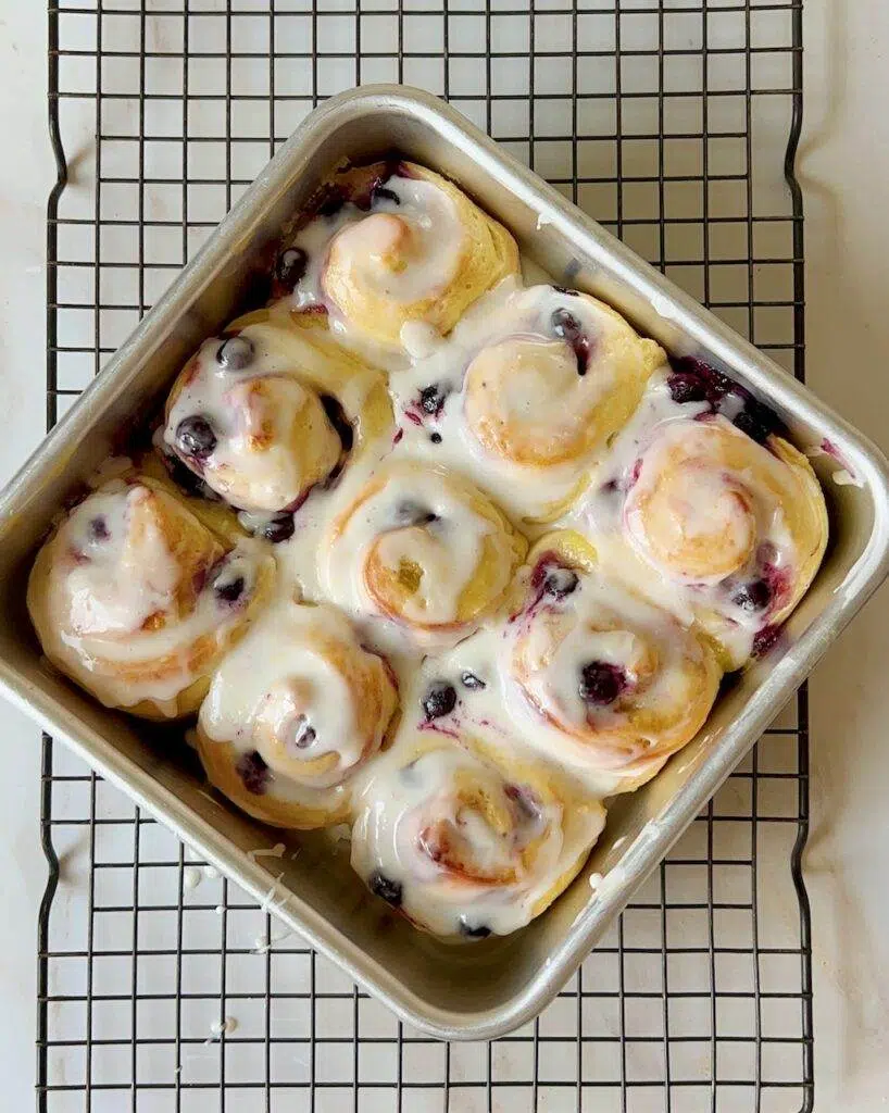 A square metal pan with iced lemon blueberry rolls, on a wire cooling rack.