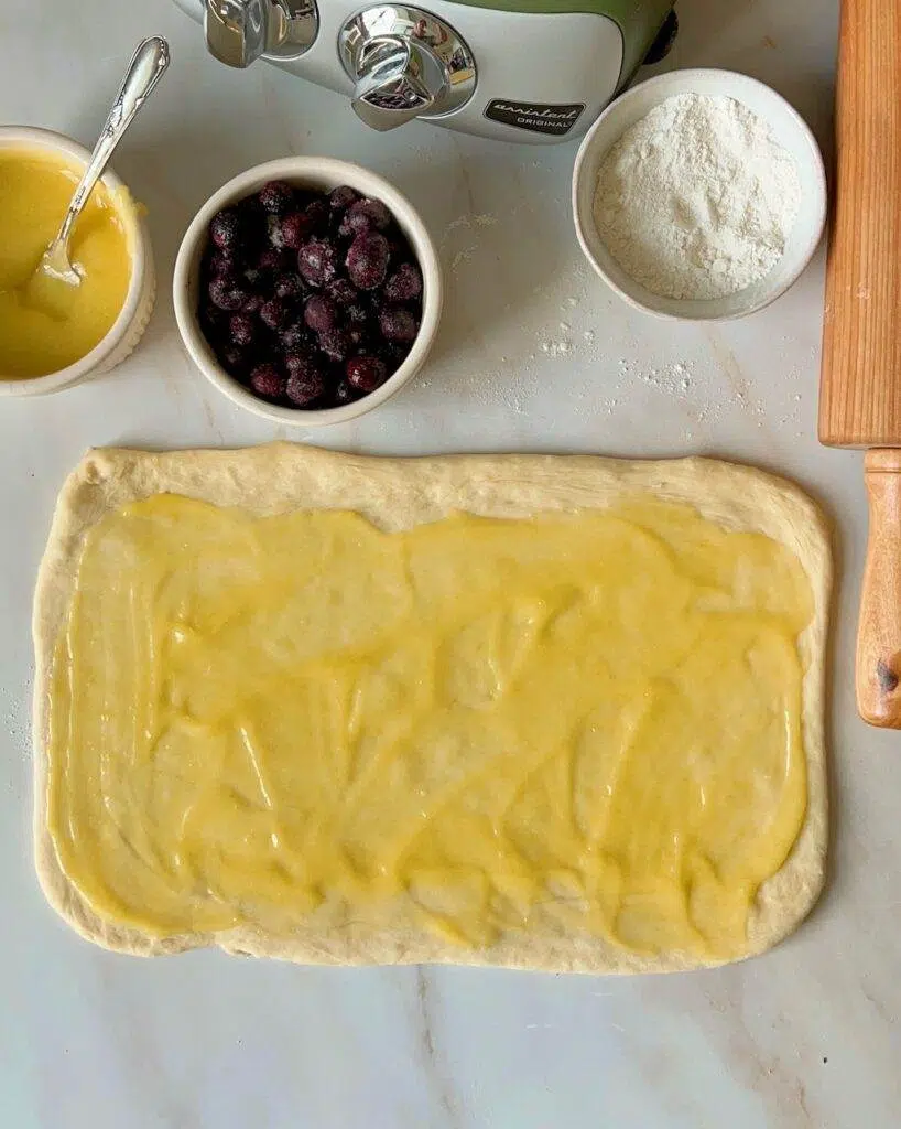 Sweet roll dough rolled out to a rectangle, with lemon curd spread over it, a bowl of lemon curd, blueberries, flour, and a rolling pin.