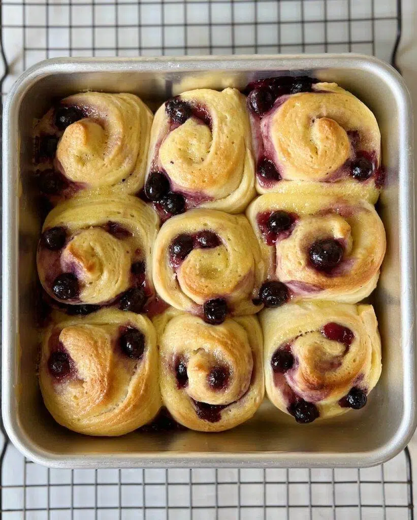 A metal baking pan with 9 baked lemon blueberry sweet rolls, resting on a wire cooling rack.