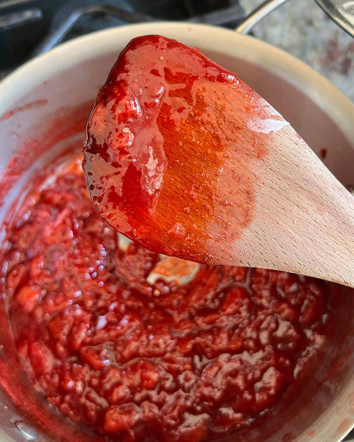 Strawberry compote, in a saucepan, with a wooden spoon over it, with strawberry compote sticking to the spoon.